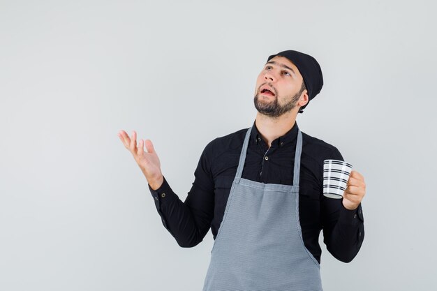 Young man in shirt, apron holding cup of drink and looking confused , front view.