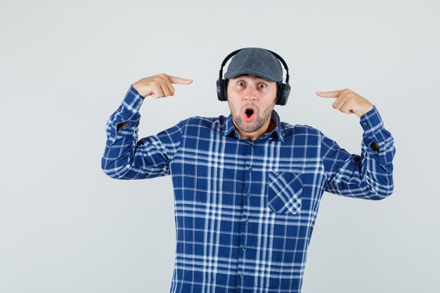 Young man in shirt, cap pointing at his headphones and looking surprised , front view.