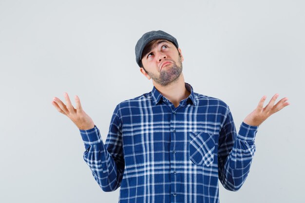 Young man in shirt, cap showing helpless gesture and looking confused , front view.