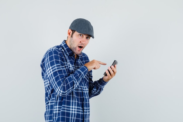 Young man in shirt, cap using calculator and looking hesitant , front view.