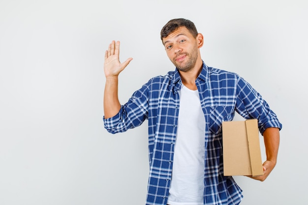 Free Photo young man in shirt showing palm while holding cardboard box and looking cheery , front view.