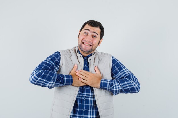 Young man in shirt,sleeveless jacket holding hands on his chest and looking excited , front view.