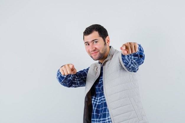 Young man in shirt,sleeveless jacket pointing forward and looking joyful , front view.