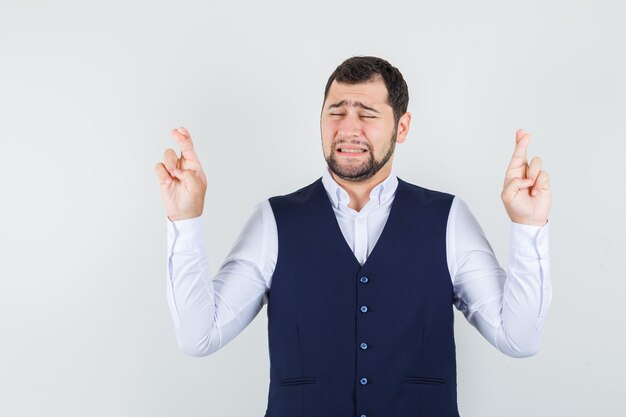 Young man in shirt, vest keeping fingers crossed and looking hopeful