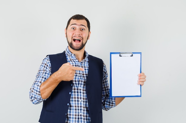 Young man in shirt, vest pointing at clipboard and looking merry , front view.