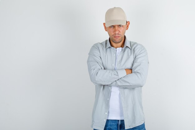 Young man in shirt with cap, jeans standing with crossed arms and looking resentful