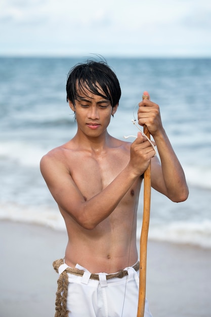 Young man shirtless on the beach with wooden bow preparing to practice capoeira