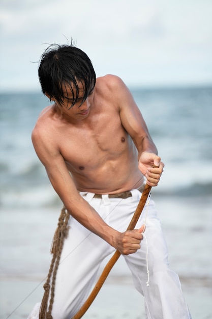 Young man shirtless on the beach with wooden bow preparing to practice capoeira