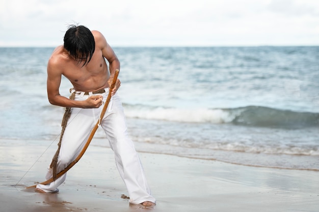 Free photo young man shirtless on the beach with wooden bow preparing to practice capoeira