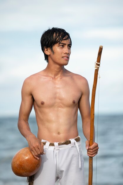 Young man shirtless on the beach with wooden bow preparing to practice capoeira