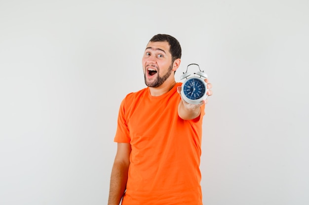 Young man showing alarm clock in orange t-shirt and looking happy. front view.