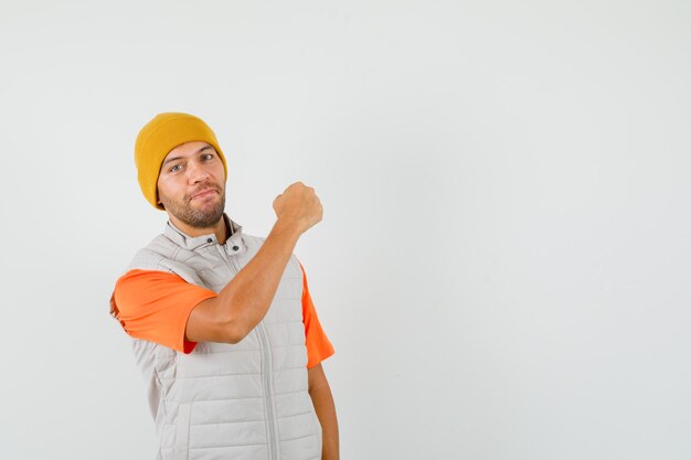 Young man showing clenched fist in t-shirt, jacket, hat and looking confident