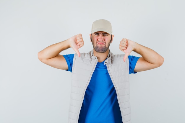 Free Photo young man showing double thumbs down in t-shirt, jacket, cap and looking sad. front view.