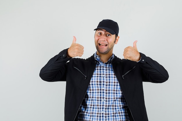 Free photo young man showing double thumbs up in shirt, jacket, cap and looking glad , front view.