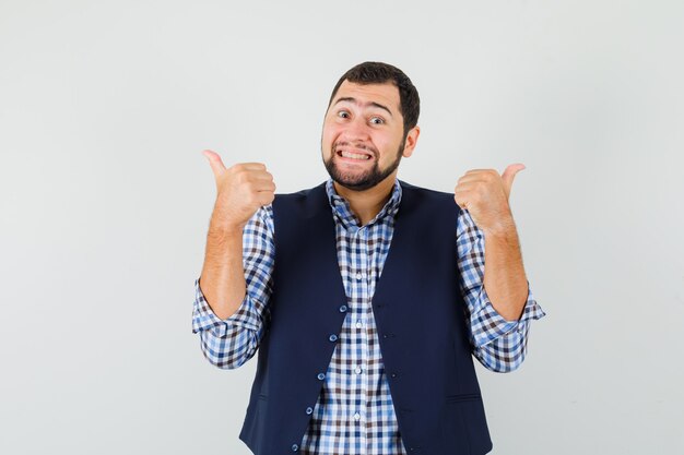 Young man showing double thumbs up in shirt, vest and looking blissful.