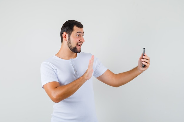 Young man showing greeting gesture while making video call in white t-shirt and looking happy .