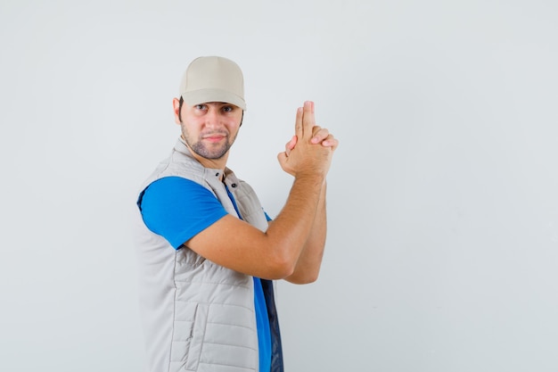 Young man showing gun gesture in t-shirt, jacket, cap and looking confident , front view.