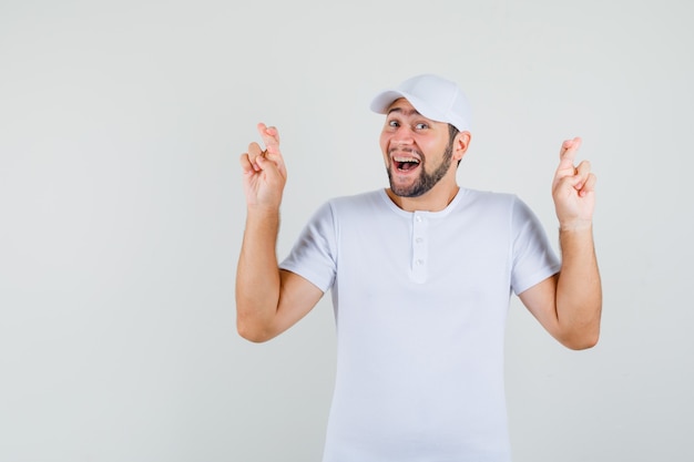 Young man showing his crossed fingers in white t-shirt,cap and looking jolly. front view.