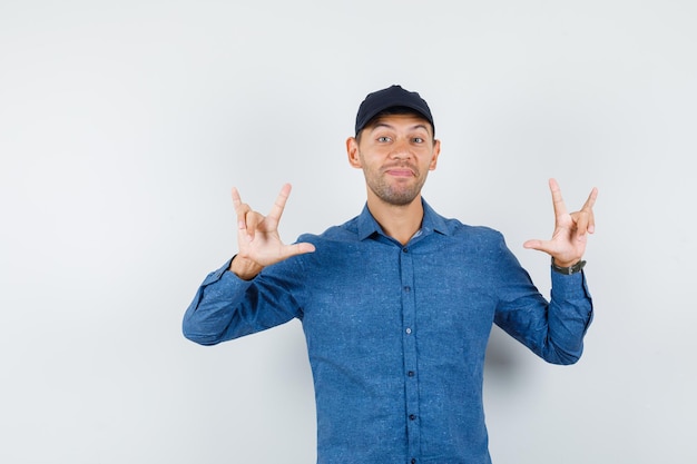 Young man showing i love you gesture in blue shirt, cap and looking cheerful , front view.