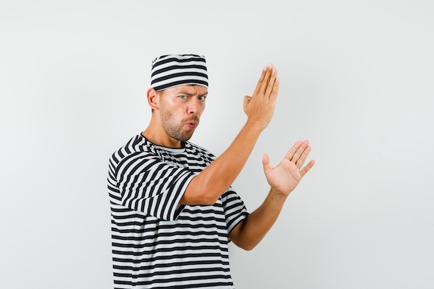 Free photo young man showing karate chop gesture in striped t-shirt, hat and looking powerful.