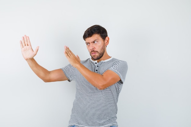 Young man showing karate chop gesture in striped t-shirt and looking angry. front view.