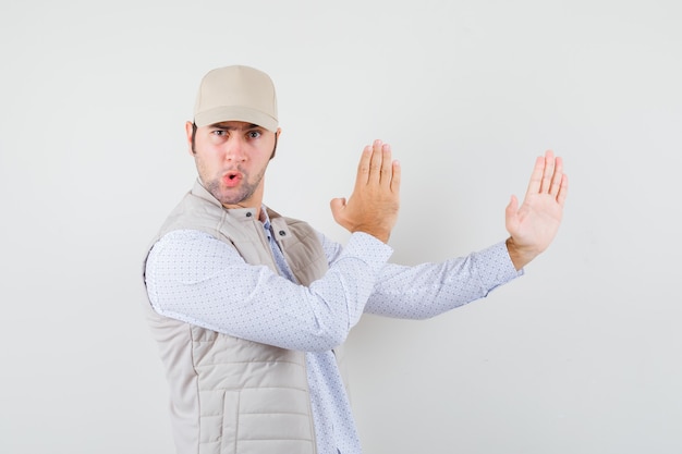 Young man showing karate chop in shirt,sleeveless jacket,cap and looking flexible. front view.