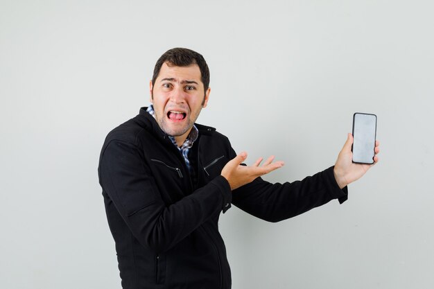 Young man showing mobile phone in shirt, jacket and looking puzzled , front view.