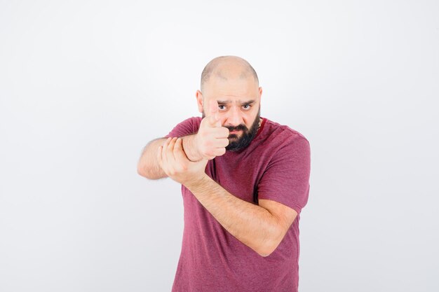 Young man showing pistol gesture in pink t-shirt and looking focused. front view.