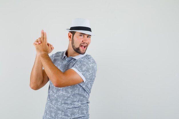 Young man showing pistol gesture in t-shirt,hat and looking lionhearted , front view. space for text