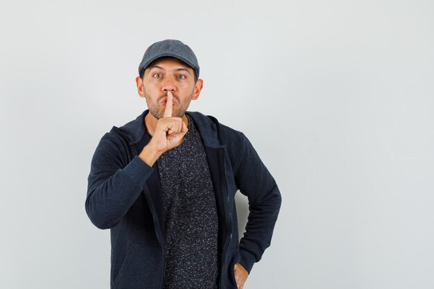 Young man showing silence gesture in t-shirt, jacket, cap and looking careful