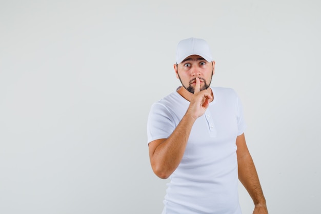 Young man showing silence gesture in white t-shirt,cap and looking focused. front view. space for text