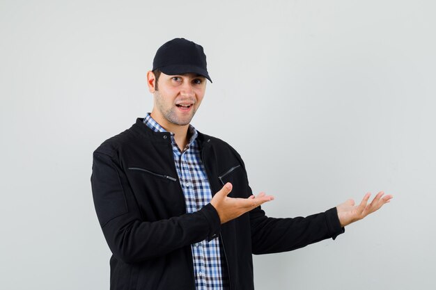 Young man showing something or welcoming in shirt, jacket, cap and looking gentle , front view.