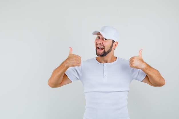 Young man showing thumb up in white t-shirt,cap and looking cheerful , front view.