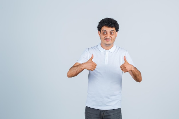 Young man showing thumbs up with both hands in white t-shirt and jeans and looking happy. front view.