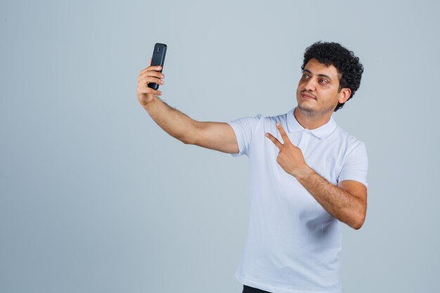 Young man showing V-sign while taking selfie in white t-shirt and looking proud , front view.