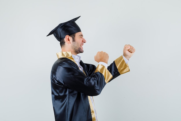 Young man showing winner gesture in graduate uniform and looking lucky. .