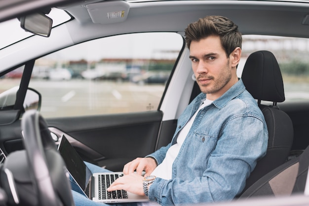 Free photo young man sitting inside the modern car with laptop looking at camera