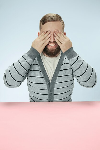 Young man sitting at table at studio with eyes closed isolated on blue. I can not see anything concept. Minimalism style