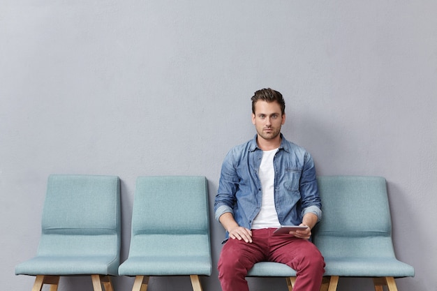 Free photo young man sitting in waiting room holding tablet