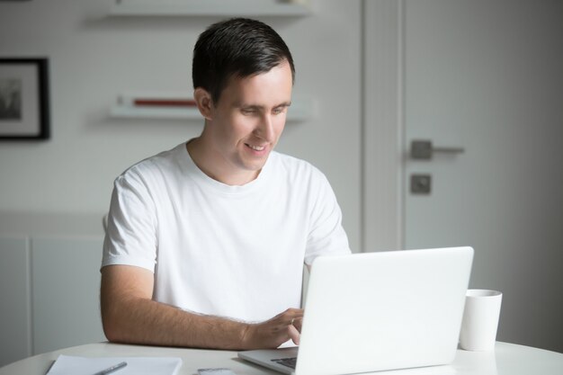 Young man sitting at the white desk working with laptop