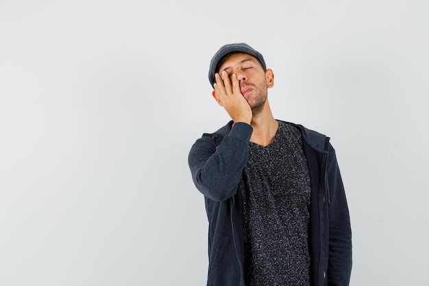 Young man sleeping with hand on cheek in t-shirt, jacket, cap and looking exhausted