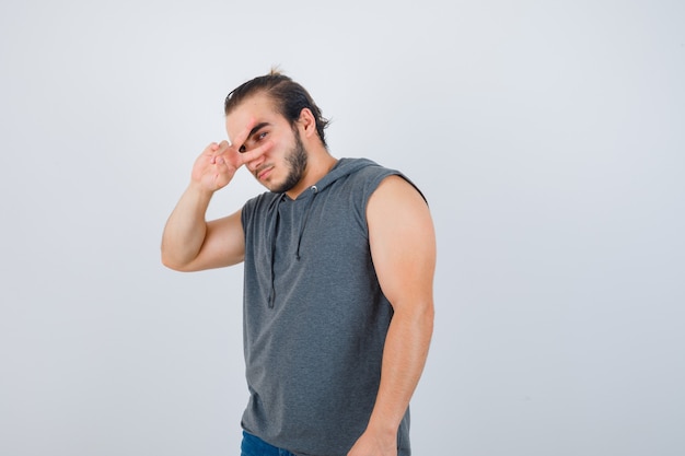 Young man in sleeveless hoodie showing V-sign on eye and looking serious , front view.