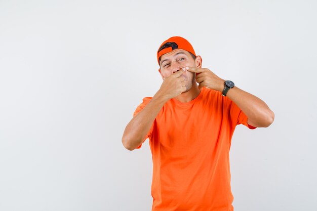 Young man squeezing her pimple on cheek in orange t-shirt and cap