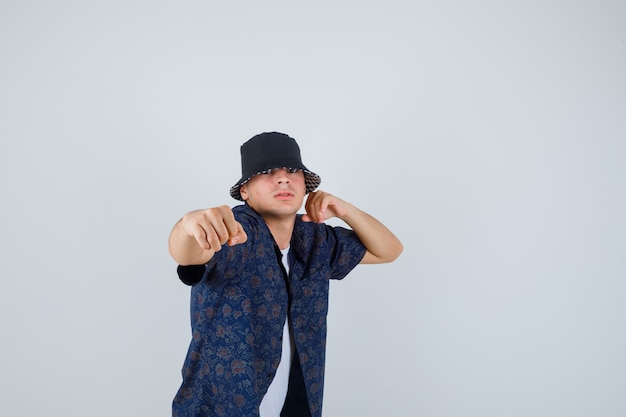 Free photo young man standing in fight pose in white t-shirt, floral shirt, cap and looking confident. front view.