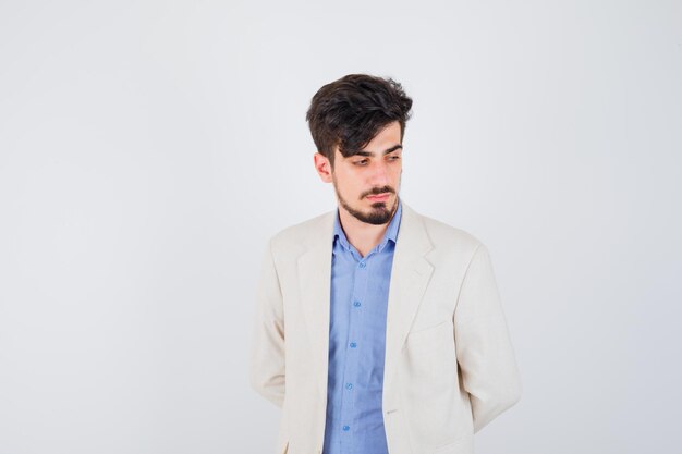 Young man standing straight and posing at front in blue t-shirt and white suit jacket and looking serious