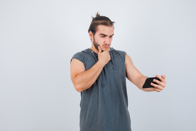 Young man standing in thinking pose, holding phone in hooded t-shirt and looking sensible , front view.