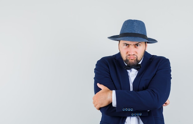 Young man standing with crossed arms in suit, hat and looking furious , front view.