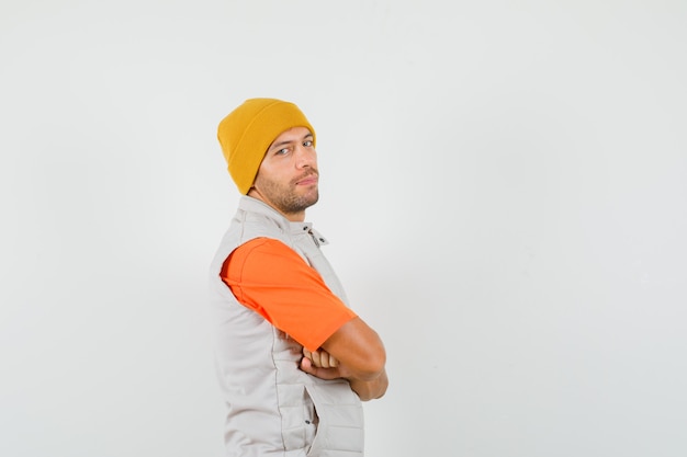 Free photo young man standing with crossed arms in t-shirt, jacket, hat and looking confident .