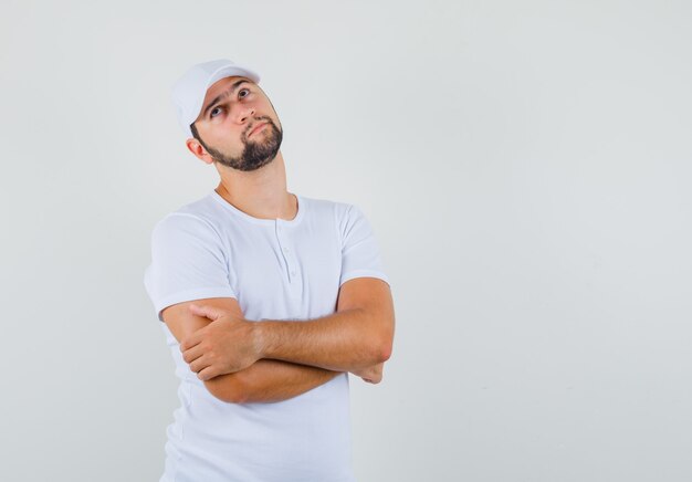 Young man standing with crossed arms in white t-shirt,cap and looking thoughtful , front view. space for text