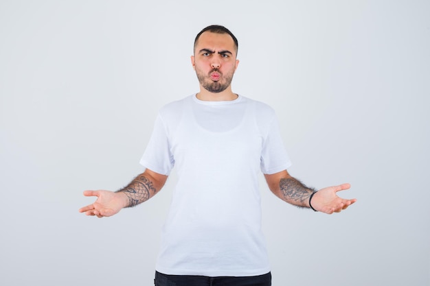 Young man stretching hands in questioning manner in white t-shirt and black pants and looking perplexed